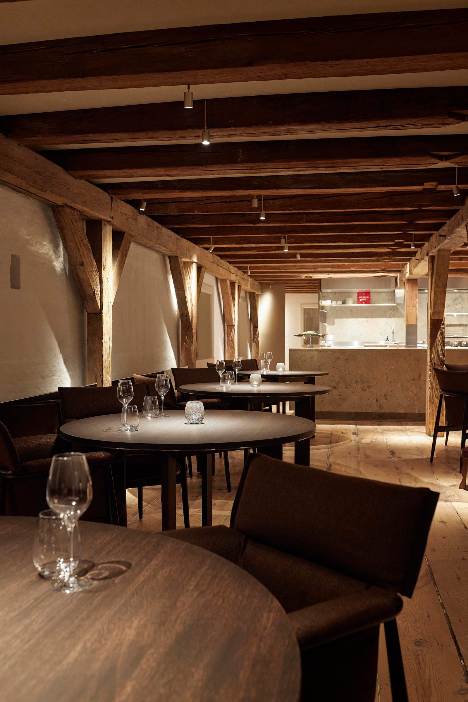 Round tables along the timber-framed dining room, with the open kitchen at the far end