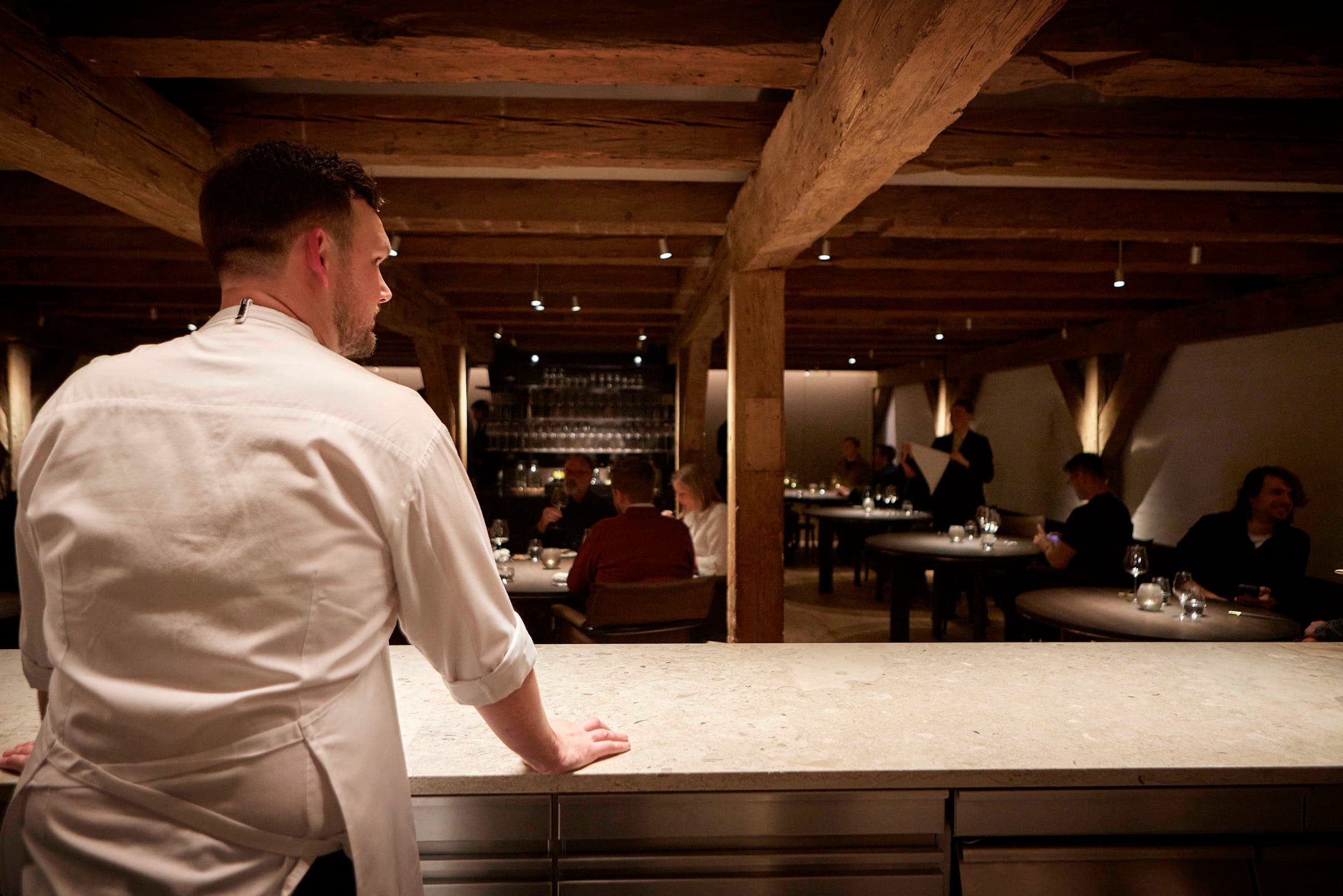 A chef at the open kitchen counter, looking out over guests in the dining room